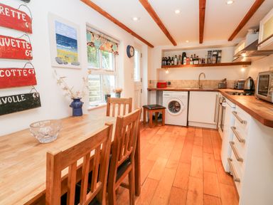 A kitchen with wooden dining table and chairs washing machine oven and shelves with bottles at Honeysuckle Cottage in St Agnes