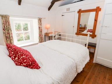 A bedroom with a white metal bed frame red patterned pillows a wooden floor a window with floral curtains a wooden framed mirror and a small desk with a lamp at Honeysuckle Cottage in St Agnes