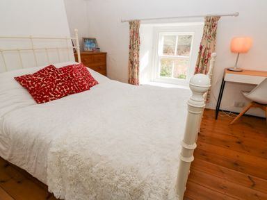 A bedroom with a white metal bed frame and red pillows near a window with floral curtains at Honeysuckle Cottage in St Agnes