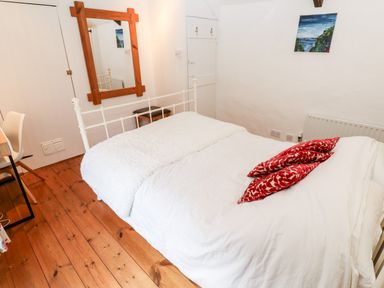 A bedroom with a white metal bed frame white bedding red patterned pillows wooden floor a wooden framed mirror and a small painting on the wall at Honeysuckle Cottage in St Agnes