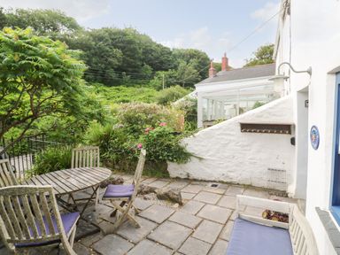 An outdoor patio with wooden table chairs and bench near a white building with greenery at Honeysuckle Cottage in St Agnes