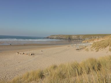 A sandy beach with grass-covered dunes a small stone building and the ocean in the background at Palm View in Holywell Bay
