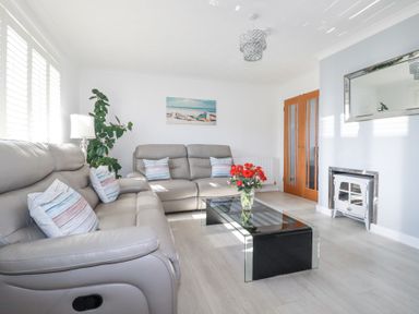 A living room with gray sofas a glass coffee table with red flowers a plant a white lamp a painting wooden doors and a white fireplace at Palm View in Holywell Bay