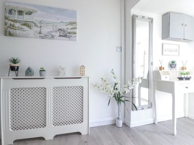 A white radiator cover with small decorative items above it and a white desk with plants and accessories beside a tall wall mirror at Palm View in Holywell Bay