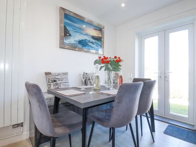 A dining area with a table set with glasses and bottles flowers on the table four grey chairs and glass doors at Palm View in Holywell Bay