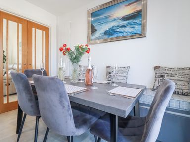 A dining area with gray chairs a table set with glasses bottles flowers and a cushioned bench with pillows at Palm View in Holywell Bay