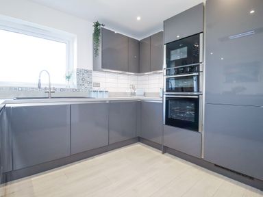 A modern kitchen with gray cabinets integrated ovens a sink under a window and tiled backsplash at Palm View in Holywell Bay