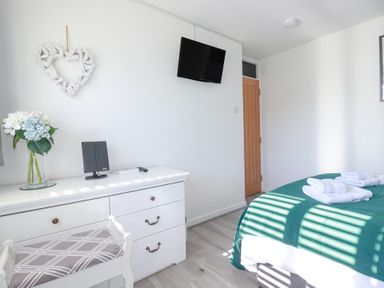 A bedroom with a white dresser a chair a wall-mounted TV and a bed with folded towels at Palm View in Holywell Bay