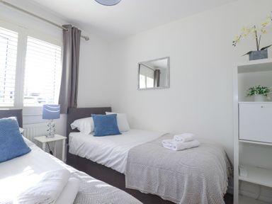 A bedroom with two single beds with blue pillows and folded towels on a shelf with plants at Palm View in Holywell Bay
