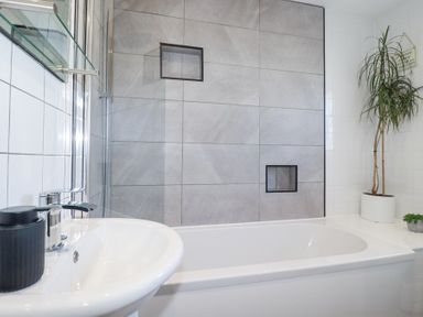 A bathroom with a bathtub a sink with a faucet a tall plant and tiled walls at Palm View Holywell Bay