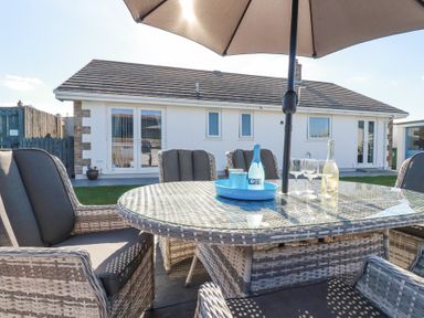 An outdoor patio with wicker chairs and glass table under an umbrella in front of a white house at Palm View in Holywell Bay