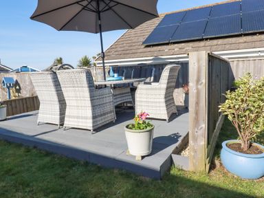 An outdoor patio with wicker chairs a table and a large umbrella solar panels on roof wooden fence and potted plants at Palm View in Holywell Bay