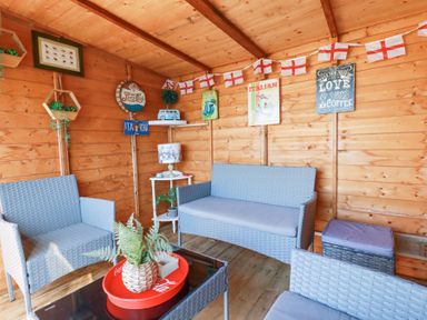 A wooden room with wicker chairs and a couch a small table with a lamp and plant a coffee table with a tray and plant and decorations on the walls at Palm View in Holywell Bay