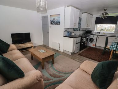 A living area with beige sofas green cushions a wooden coffee table a TV on a stand and an open kitchen with white cabinets and appliances at 13 Gower Holiday Village in Scurlage