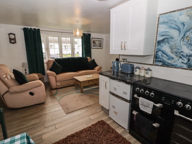 A combined kitchen and living room with beige sofas a coffee table a black stove white cabinets and a window with dark curtains at 13 Gower Holiday Village Scurlage
