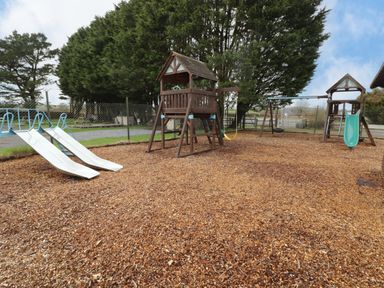 An outdoor playground with two white slides a wooden play structure a swing set and wood chip ground covering at 13 Gower Holiday Village in Scurlage