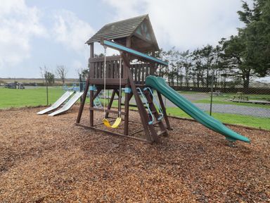 An outdoor playground with a wooden play structure featuring a slide swings and ladders at 13 Gower Holiday Village in Scurlage