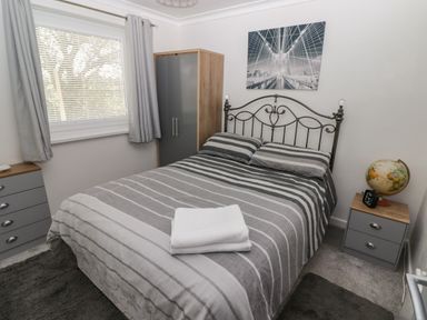 A bedroom with a double bed striped bedding a wardrobe two bedside tables a window with curtains and a wall picture at 13 Gower Holiday Village in Scurlage