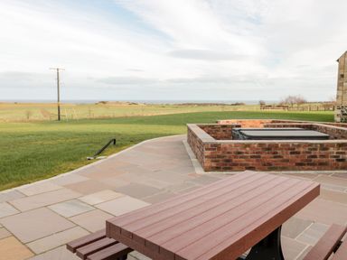 An outdoor patio with a brown picnic table and a sunken hot tub surrounded by a brick wall with green fields and the sea in the background at Glebe Cottage in Embleton