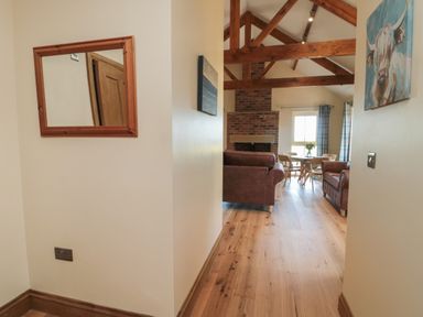 A hallway with a mirror leading to a living room with wooden beams a brick fireplace brown sofas and a dining table at Glebe Cottage in Embleton