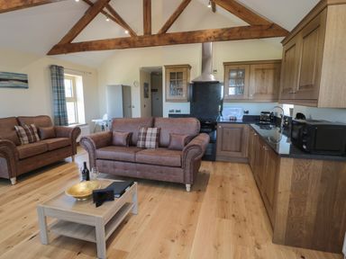 A living room and kitchen area with two brown sofas a wooden coffee table with a bottle and glass a kitchen with wooden cabinets and appliances at Glebe Cottage in Embleton