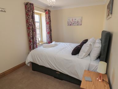 A bedroom with a bed white bedding pillows a window with patterned curtains and a wooden nightstand with a lamp at Glebe Cottage in Embleton