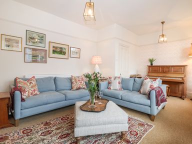 A living room with two blue sofas a gray ottoman a piano and framed pictures on the wall at Collett House in Southwold