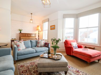A living room with blue sofas a red chaise lounge a piano and a coffee table with flowers and wine bottle at Collett House in Southwold