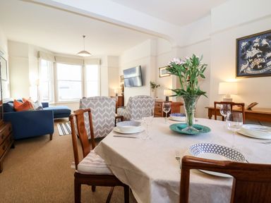 A dining area with a set table and a vase of flowers next to a living room with chairs a blue sofa and a wall-mounted TV at Collett House in Southwold