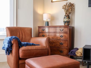 A leather armchair with a blue blanket next to a wooden chest of drawers with a lamp and vase on top with rolled blankets beside it at Collett House in Southwold
