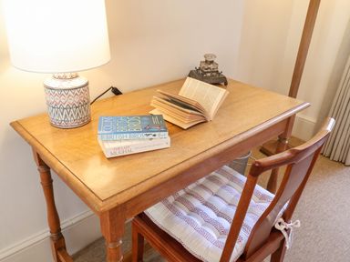 A wooden desk with a lamp, books, an open book, and a wooden chair with a cushion at Collett House in Southwold