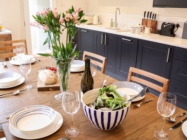 A kitchen with a wooden dining table set with plates glasses a bottle a bowl of salad and flowers at Collett House in Southwold