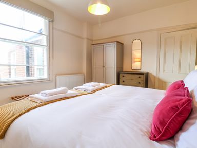 A bedroom with a large bed with red pillows and white towels on a mustard blanket a wardrobe a chest of drawers and a window at Collett House in Southwold