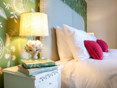 A bedside table with a lamp and books next to a bed with white pillows and pink cushions at Collett House Southwold