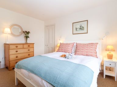A bedroom with a bed with patterned pillows and a blue blanket a wooden dresser with a round mirror and flowers and a bedside table with a lamp at Collett House in Southwold