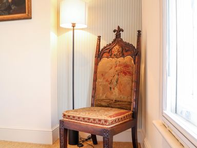 A wooden chair with an embroidered back and seat next to a floor lamp and window at Collett House in Southwold