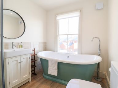 A bathroom with a green freestanding bathtub a white sink with cabinet a round mirror and a window at Collett House in Southwold