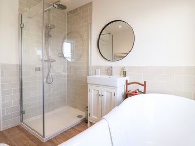 A bathroom with a glass shower enclosure a white sink under a round mirror and a wooden chair next to a white bathtub at Collett House Southwold