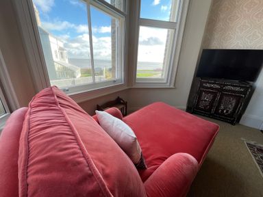 A corner with a large red sofa facing windows showing a sea view and a television on a cabinet at Collett House in Southwold
