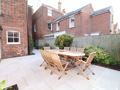 An outdoor patio area with a wooden dining table and eight chairs surrounded by plants and a wooden fence at Collett House in Southwold