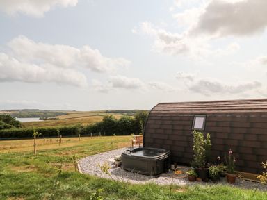 A curved roof cabin with a hot tub and potted plants in a field with rolling hills and a lake in the background at Ash in Saltash
