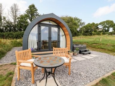 An outdoor seating area with wooden chairs and a mosaic table in front of a modern pod-style cabin with glass doors and a hot tub at Ash in Saltash