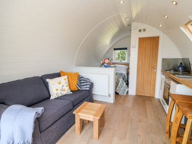 A living area with a dark sofa and cushions a small wooden table kitchenette with stools a door and a bed in a small room at Ash in Saltash