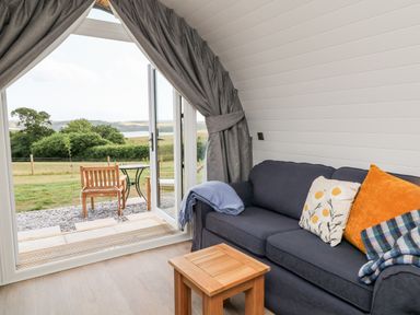 A living area with a dark sofa and cushions next to an open door leading to an outdoor seating area with wooden chairs and a table at Ash in Saltash
