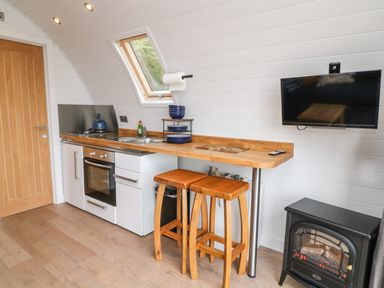 A small kitchen area with oven sink wooden counter stools and a wall mounted tv at Ash in Saltash