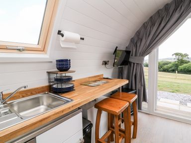 A kitchen area with a wooden countertop two wooden stools a sink stacked plates bowls a mounted television and a large window with curtains at Ash in Saltash