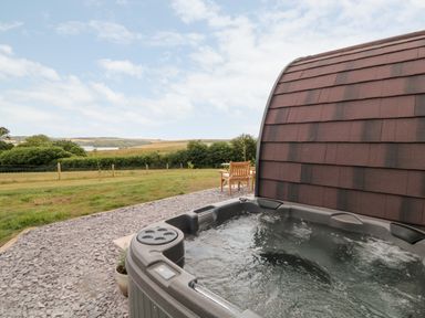 An outdoor hot tub next to a curved wooden structure with a table and chairs on a gravel area and fields in the background at Ash in Saltash