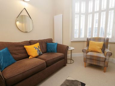 A living room with a brown sofa and cushions a patterned armchair and a round mirror at Derwent Cottage in Keswick