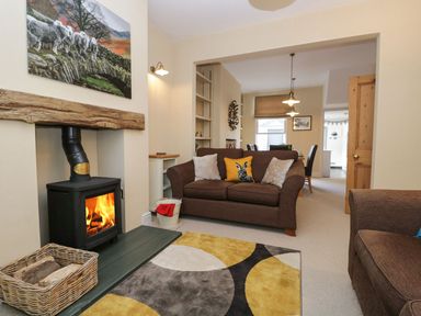A living room with a wood burning stove a brown sofa with cushions and a dining area visible in the background at Derwent Cottage in Keswick