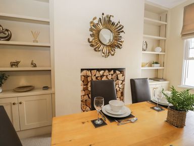 A dining area with a wooden table set with plates glasses and cutlery next to shelves and a wall with stacked firewood at Derwent Cottage Keswick